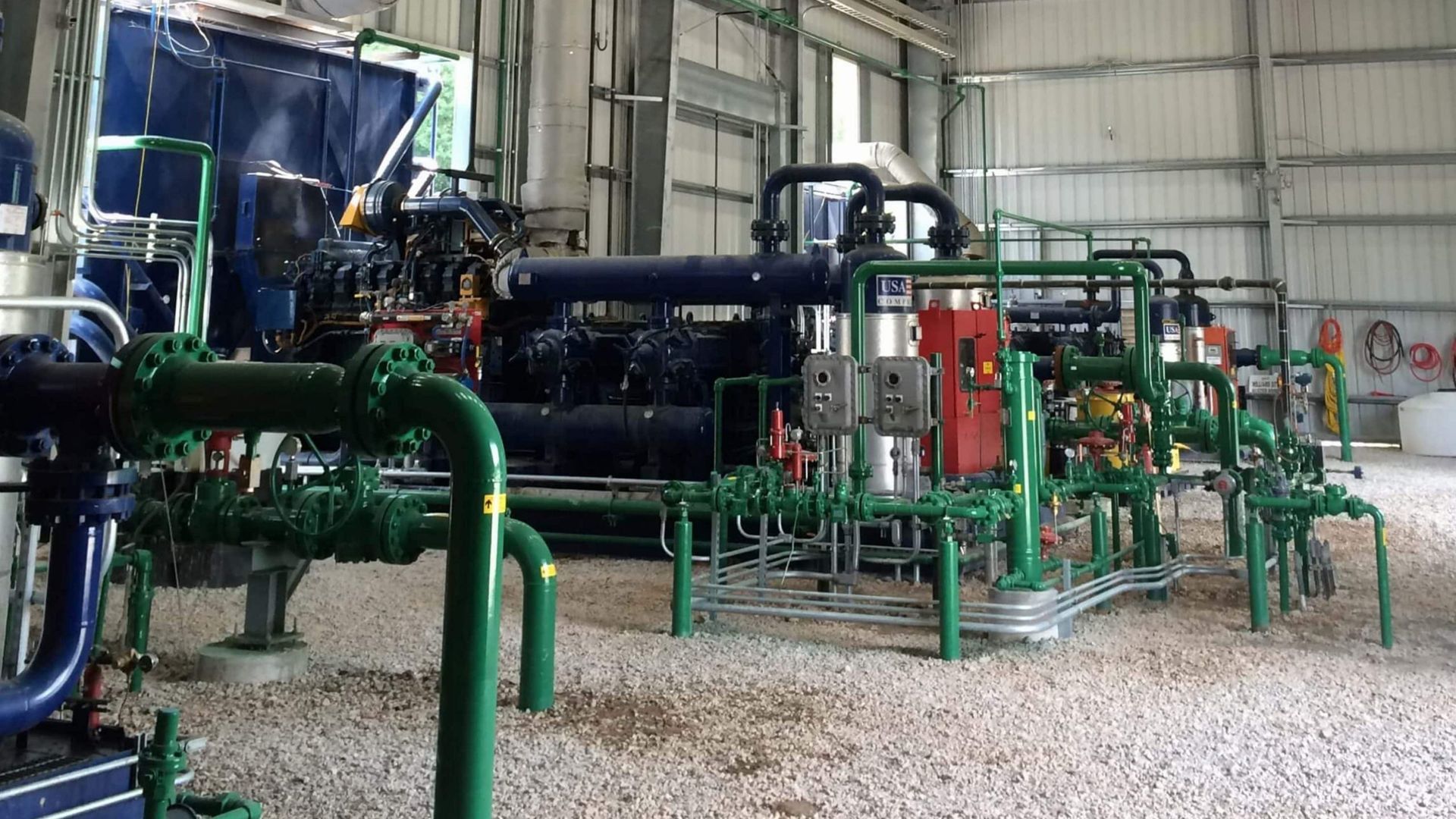 Industrial machinery and green pipes inside a factory workshop, with metal equipment and gravel floor.