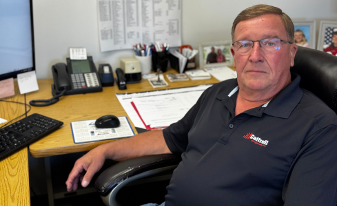 Man sitting at an office desk with a computer, phone, and paperwork, wearing a navy polo shirt.