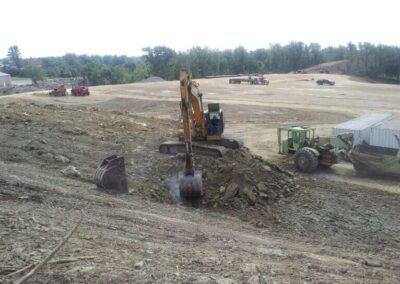 Excavator digging dirt at a construction site, with a tractor and fields in the background.