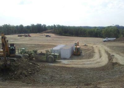 Construction site with heavy equipment, dirt roads, and a white storage tent near a tree line.