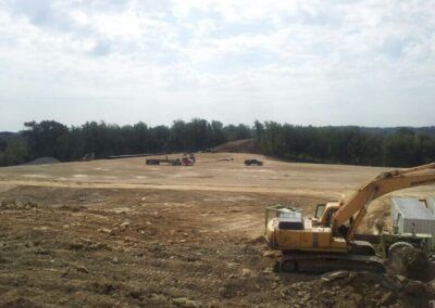 Construction site with excavator in foreground and cleared dirt field stretching to a tree line