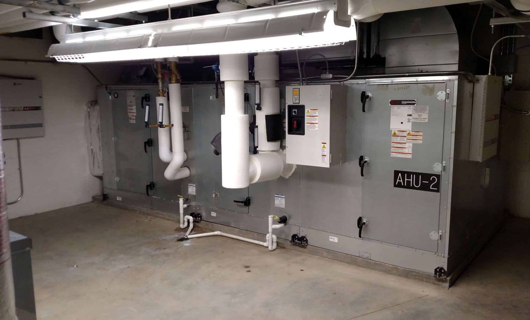 Basement HVAC machinery with ducts, pipes, and a large gray unit against a concrete wall.