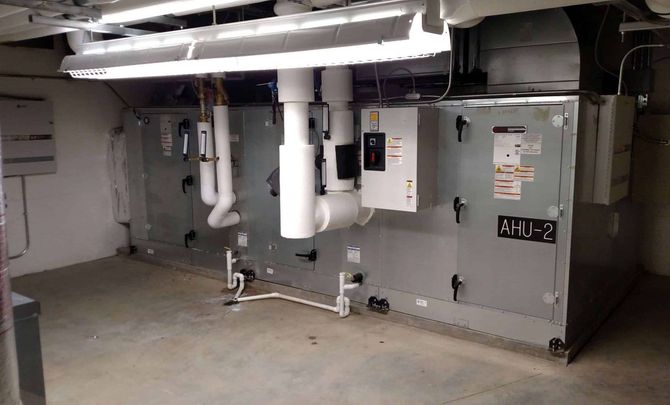 Basement HVAC machinery with ducts, pipes, and a large gray unit against a concrete wall.