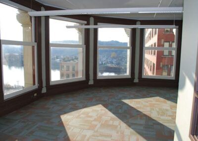 Empty room with large bay windows overlooking a city and water, carpeted floor, and red-brown trim