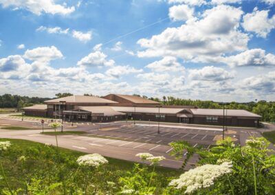 Low, single-story building with tan roof in a paved lot, viewed from across green shrubs under a cloudy blue sky