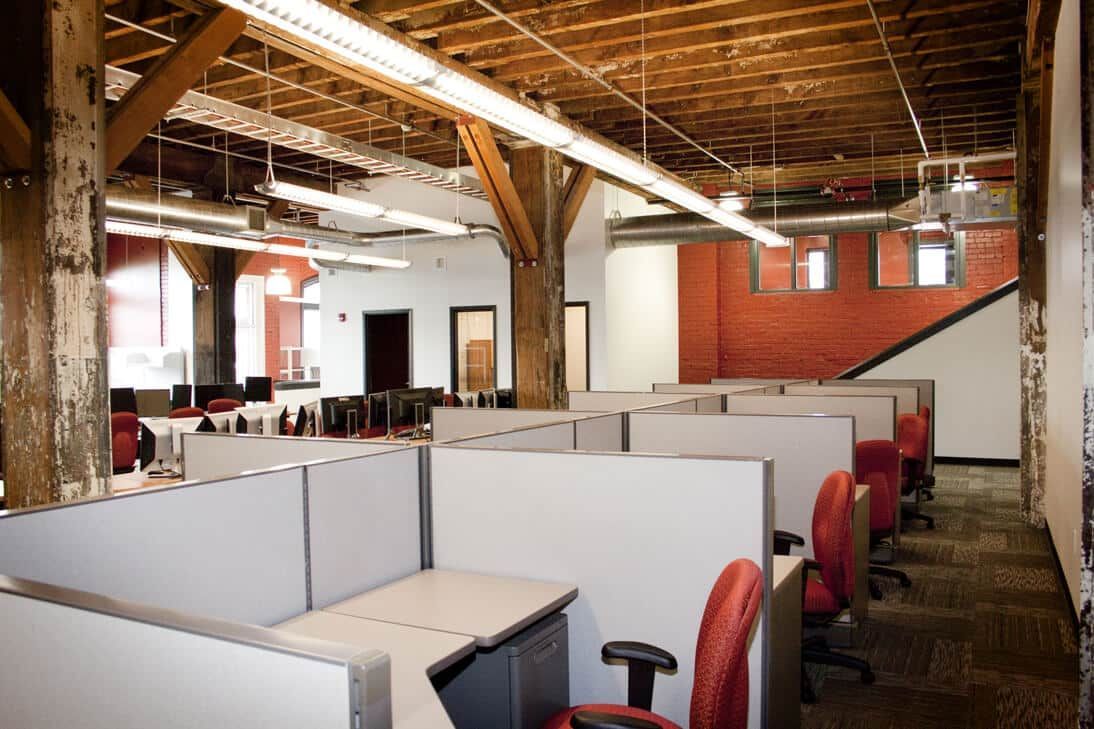 Empty office cubicles in a rustic open-plan workspace with exposed wood beams and red chairs.