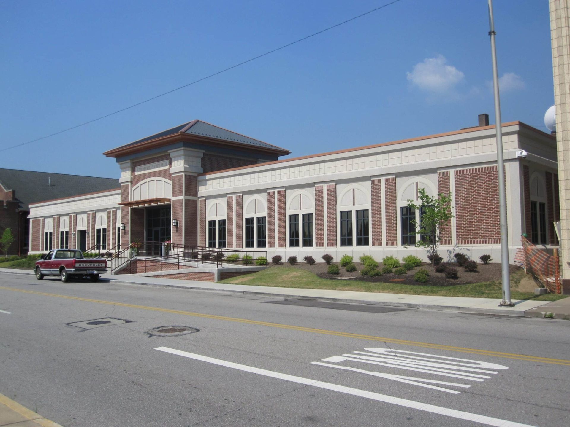 Small brick civic building with a central entrance, tall windows, and a parked car along the street.