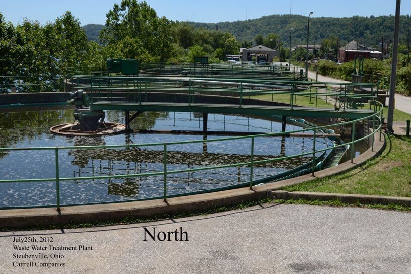 Outdoor water treatment basin with green railings, a central fountain, and hillside buildings in the background.