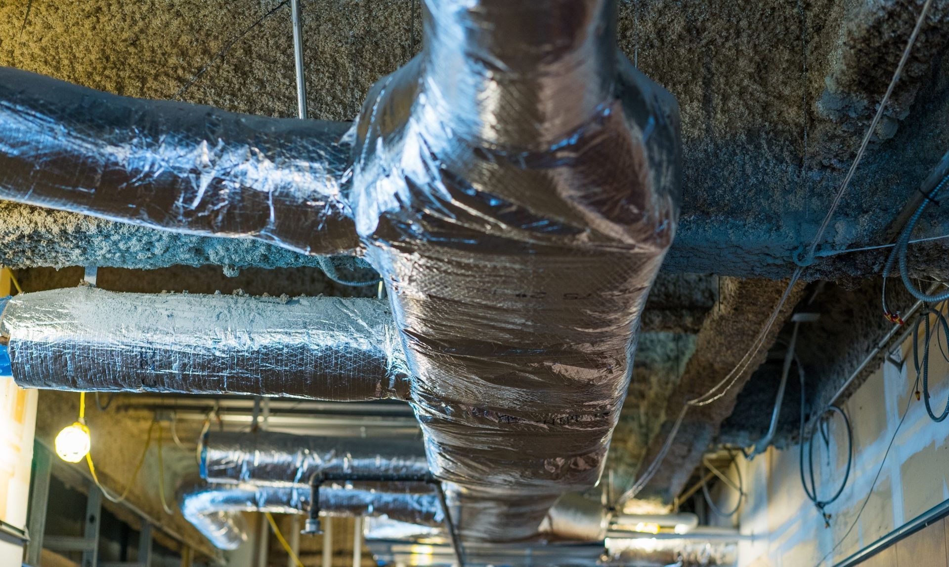 Insulated HVAC ducts and pipes running across an industrial ceiling with reflective silver wrapping.
