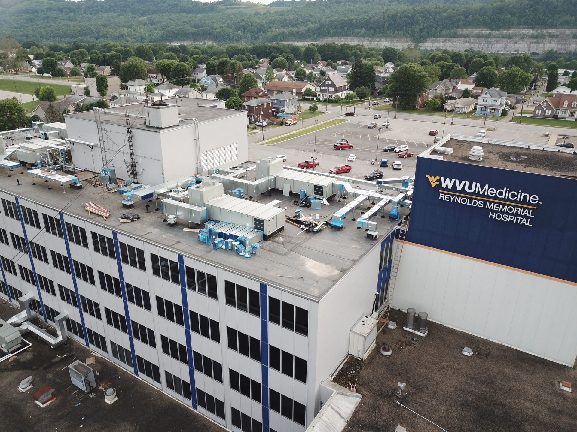 Aerial view of a white industrial building with rooftop equipment and a large parking lot near green hills.