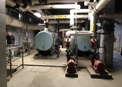 Basement mechanical room with large boilers, pipes, and red pumps lined up under low ceilings.