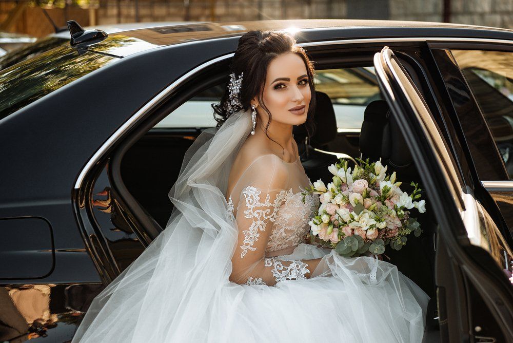 Bride in a white gown and veil holding a bouquet while seated in a black limousine.