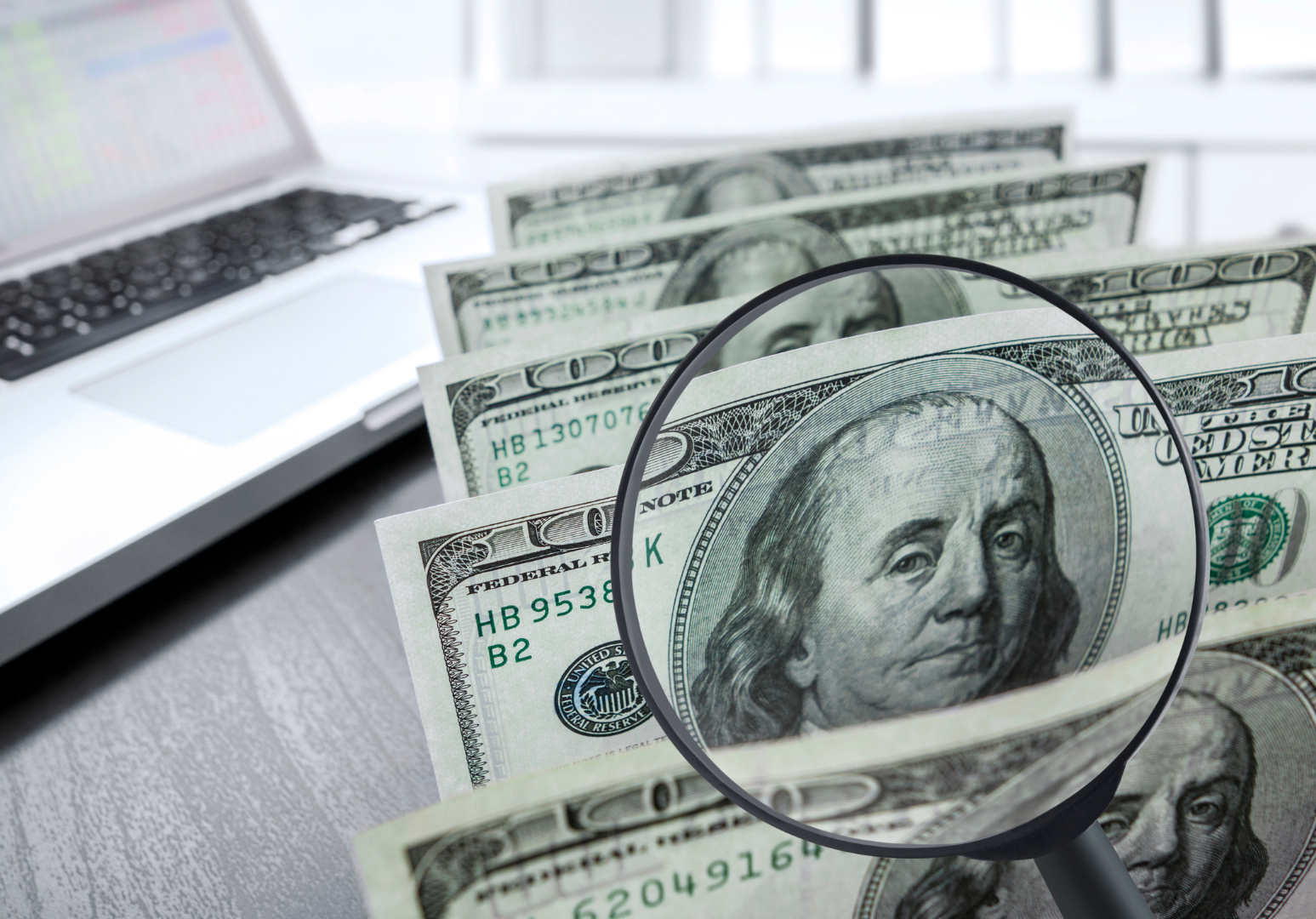 A magnifying glass is sitting on top of a pile of money next to a laptop.