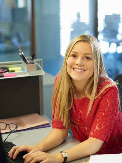 A woman in a red shirt is smiling while using a laptop