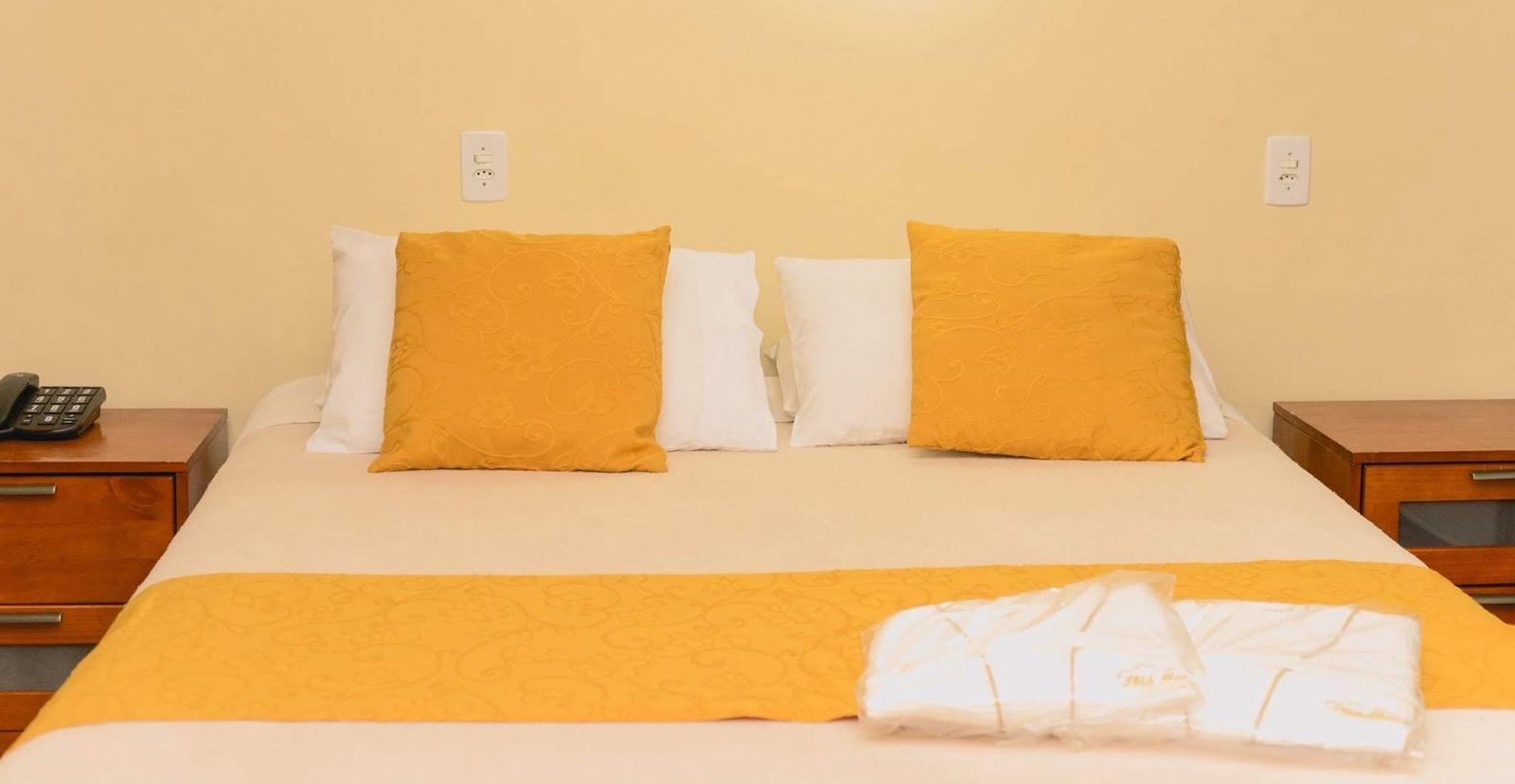 Cama de hotel com lençóis brancos, travesseiros amarelos e toalhas de mesa amarelas combinando.