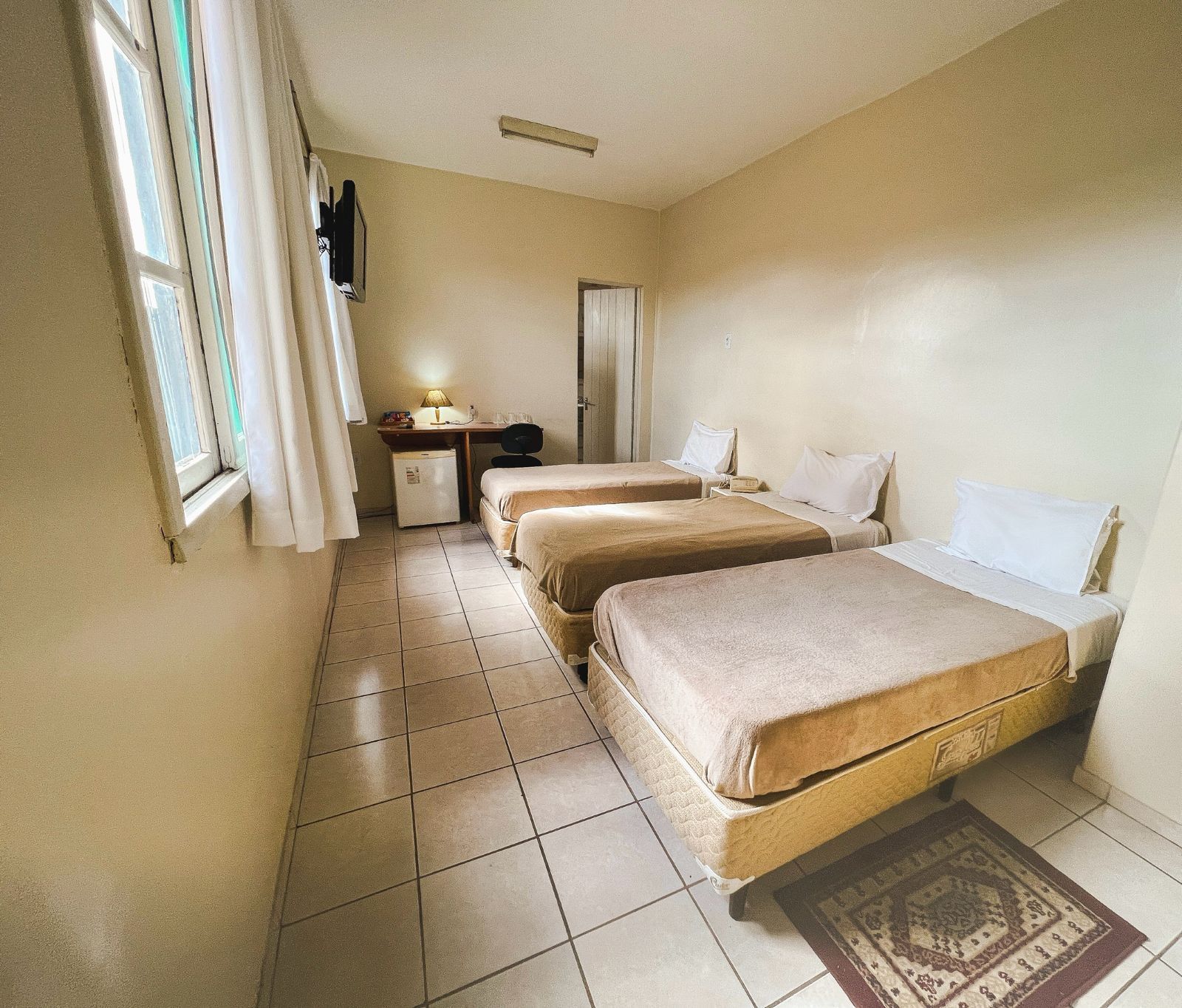 Quarto de hotel luminoso com três camas de solteiro, piso de azulejo e uma pequena escrivaninha junto à janela.