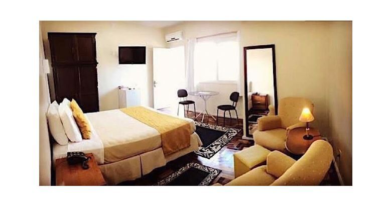 Quarto aconchegante com cama, poltrona, espelho de chão, escrivaninha e iluminação suave de abajur.