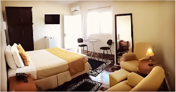 Quarto de hotel aconchegante com cama, poltrona, abajur, espelho e luz natural.