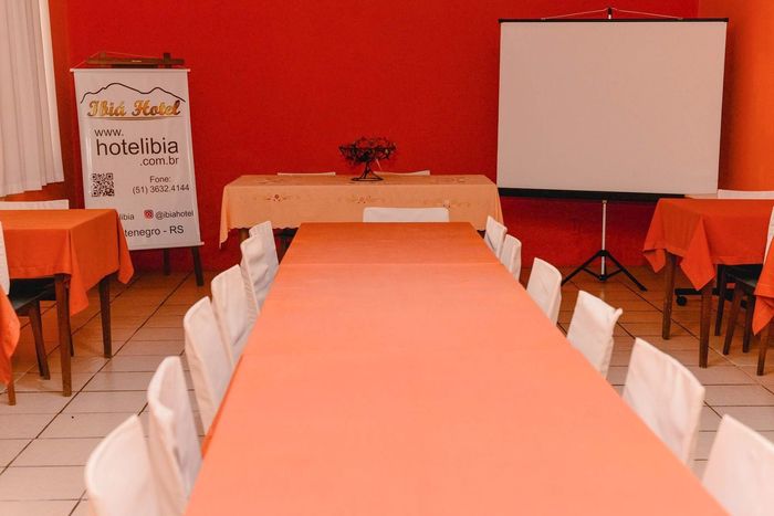 Sala de conferências com toalhas de mesa laranja, cadeiras brancas, uma tela de apresentação e uma faixa em uma parede vermelha.