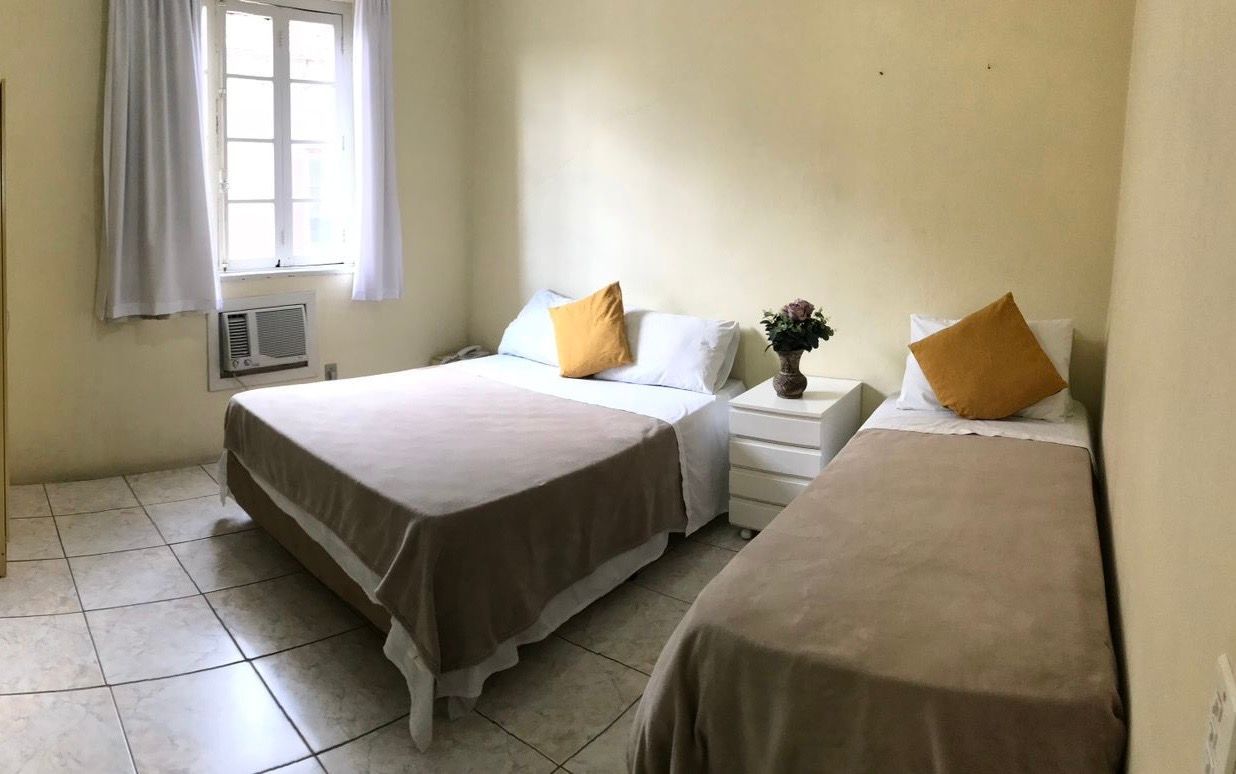 Quarto pequeno com duas camas de solteiro, paredes brancas, piso de azulejo e ar-condicionado de janela.