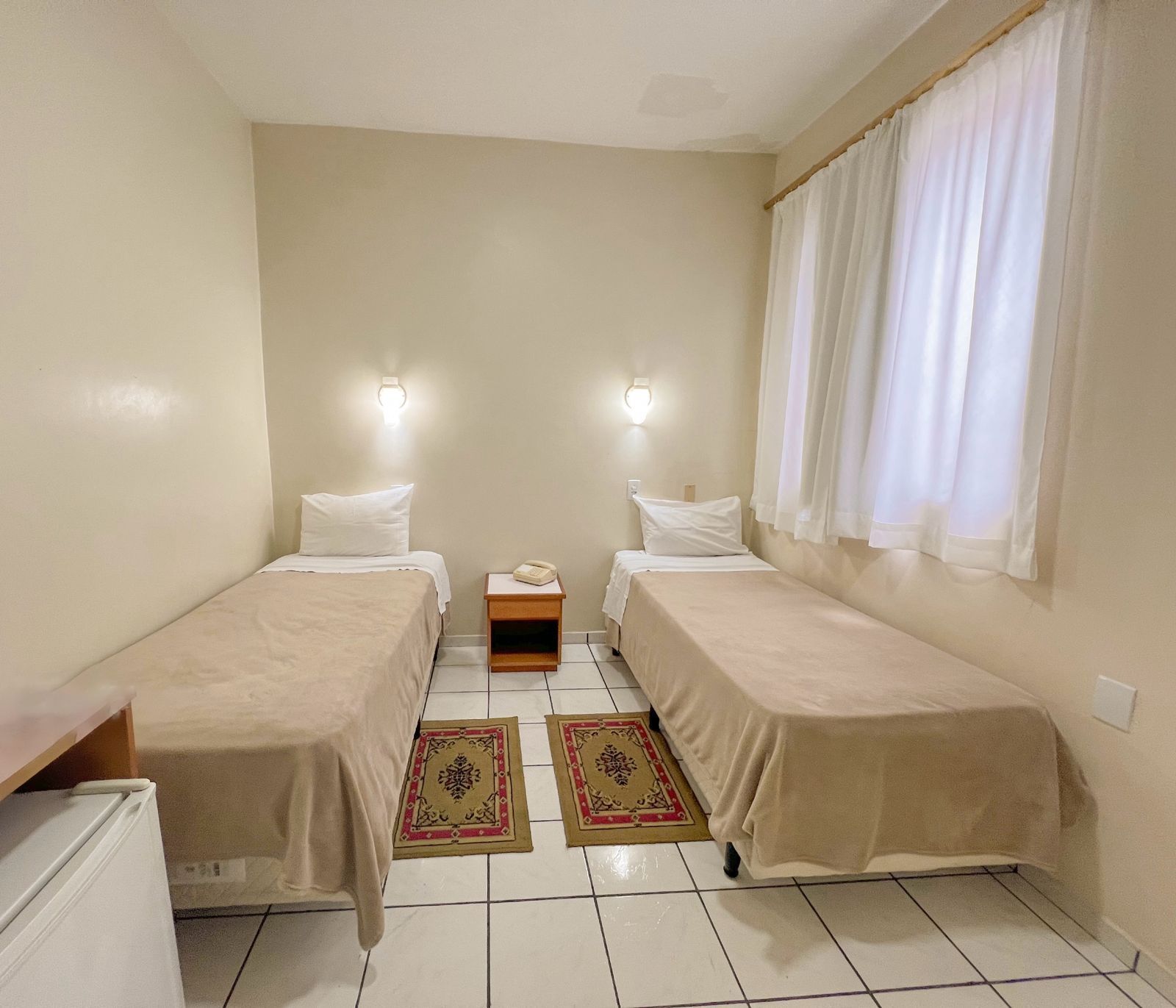 Quarto pequeno com duas camas de solteiro, cortinas brancas, paredes bege e piso de azulejo.