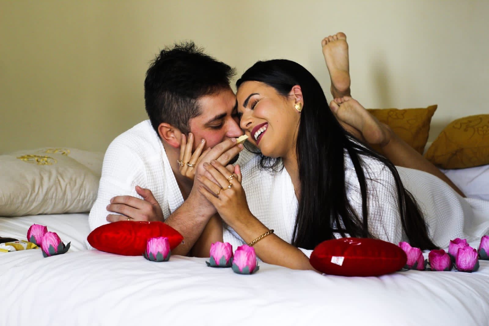 Casal sorrindo e com os rostos se tocando em uma cama com pétalas de rosas vermelhas e uma almofada em formato de coração vermelho.