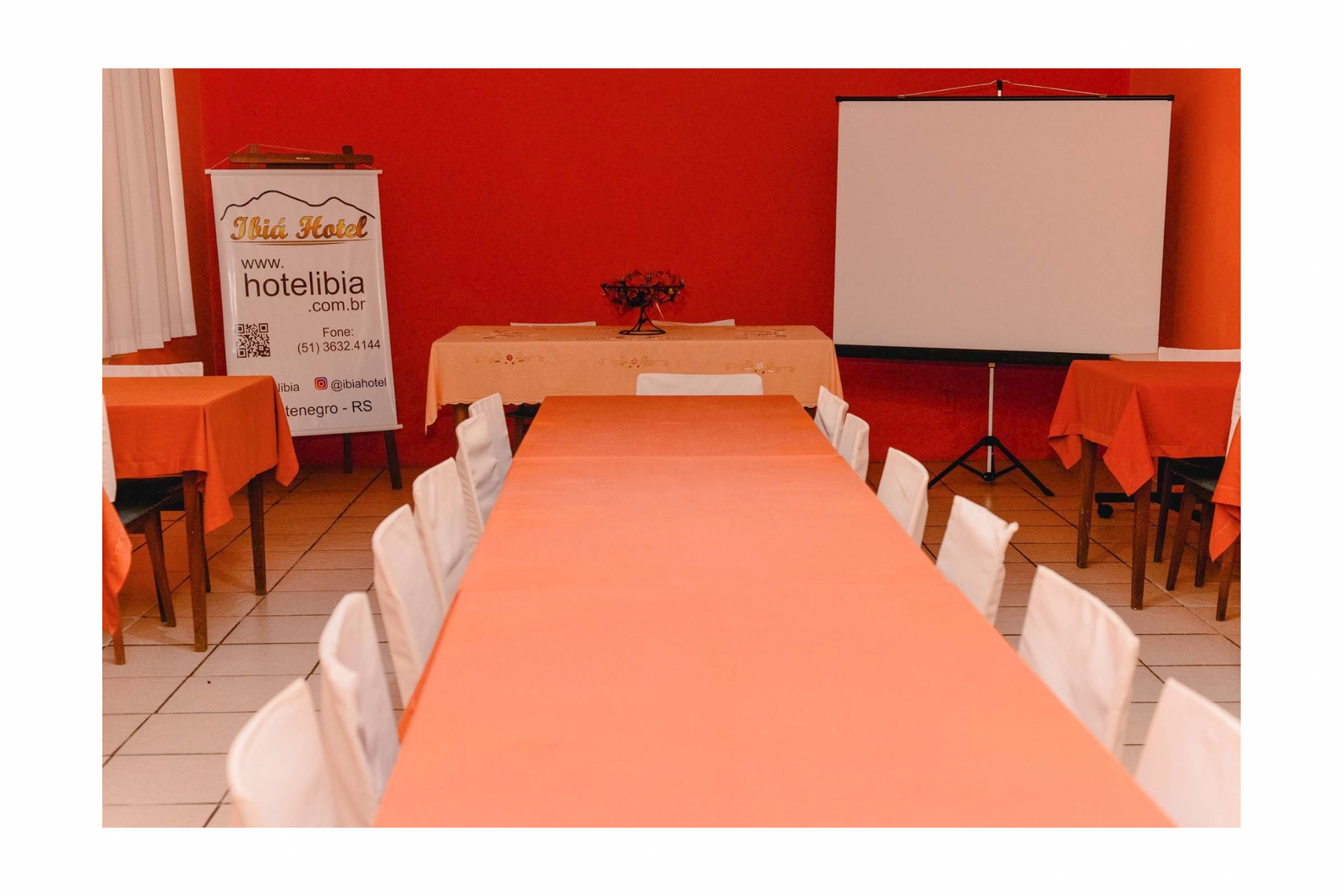 Sala de conferências com mesa comprida cor pêssego, cadeiras brancas, paredes laranja e tela de projeção.
