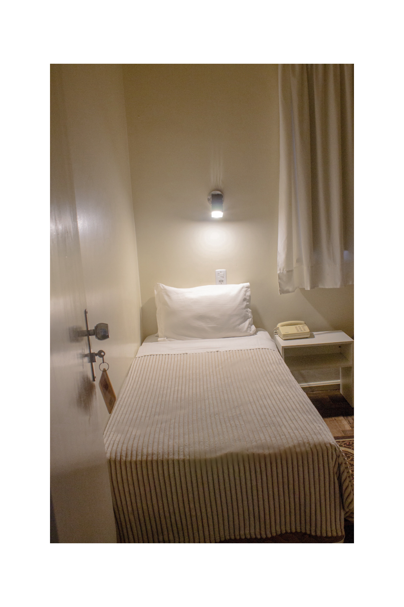 Cama de solteiro pequena em um quarto bege com um abajur de parede, cortina e telefone ao lado da cama.
