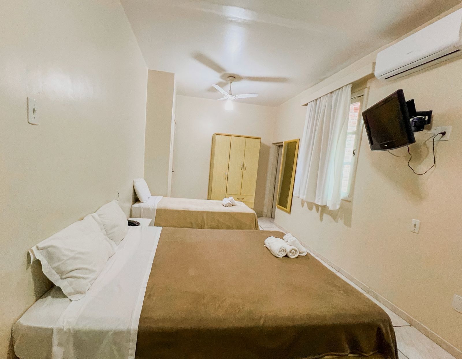 Quarto de hotel pequeno com duas camas, paredes bege, ventilador de teto, cortinas e TV de parede.