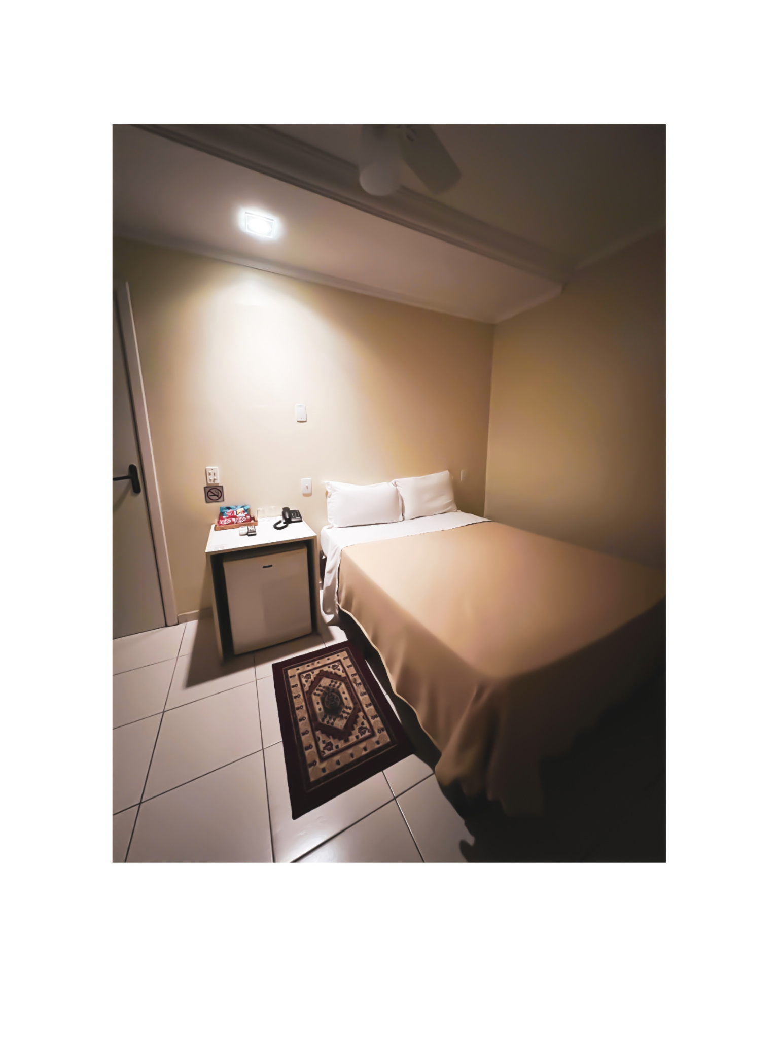 Quarto de hotel pequeno com cama arrumada, mesa de cabeceira, tapete estampado e luz de teto aconchegante.