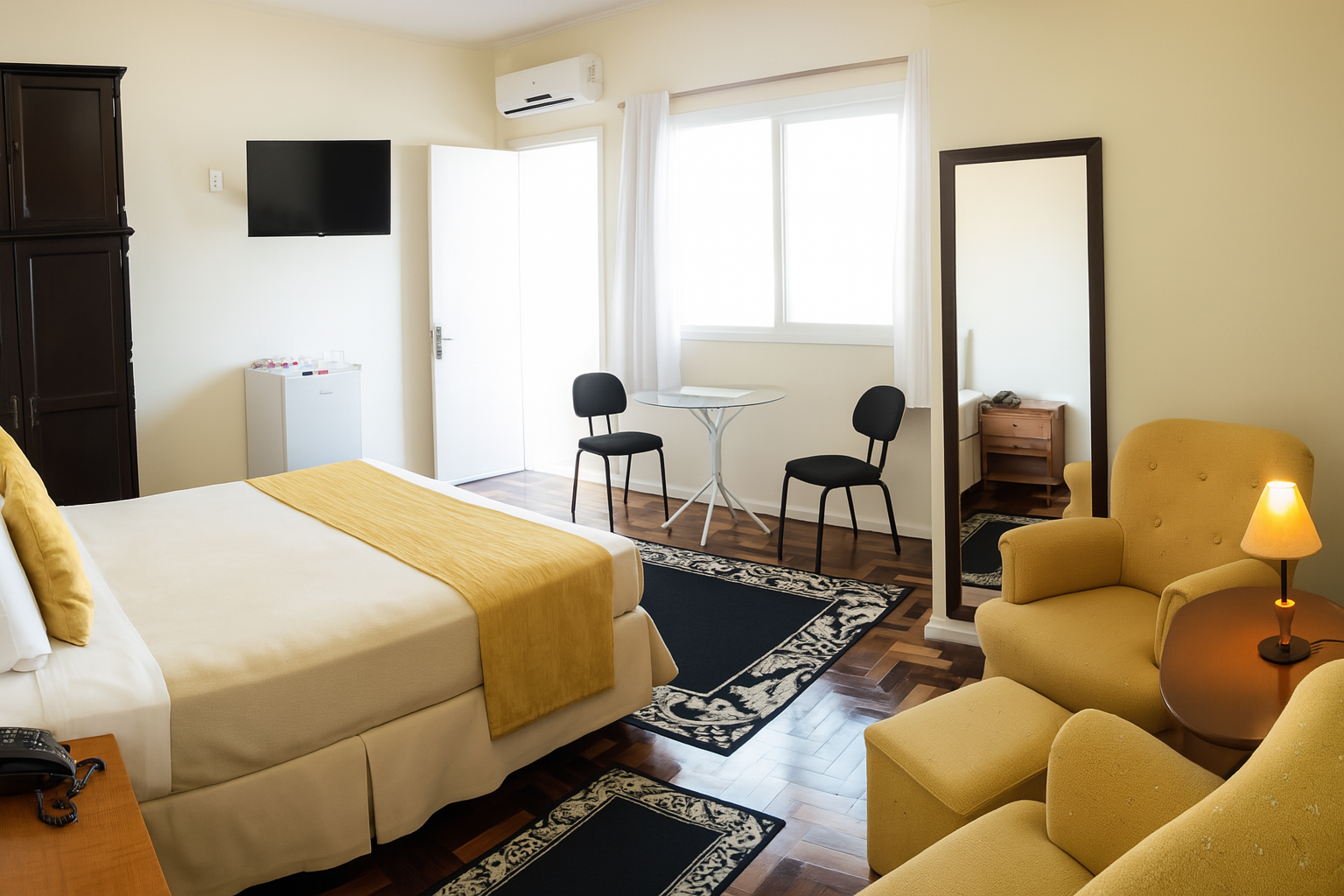 Quarto de hotel aconchegante com cama, cadeiras junto à janela, poltrona, abajur e TV de parede.