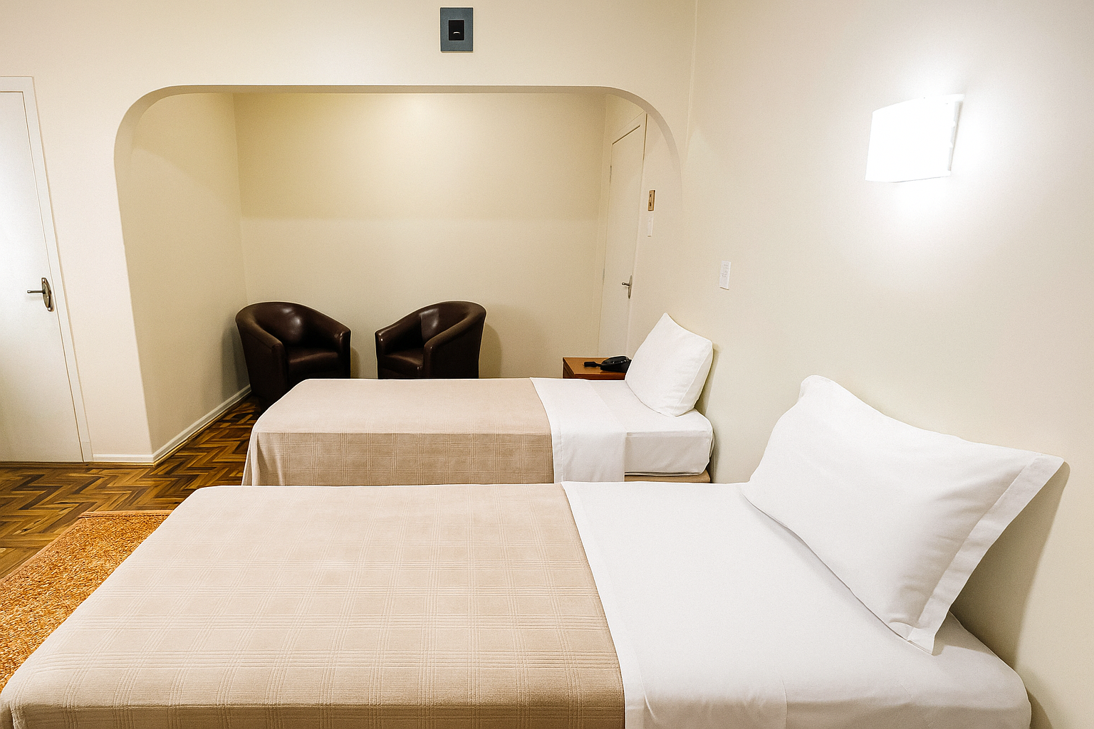 Quarto de hotel com duas camas de solteiro, paredes brancas e duas cadeiras pretas em uma área de estar rebaixada.