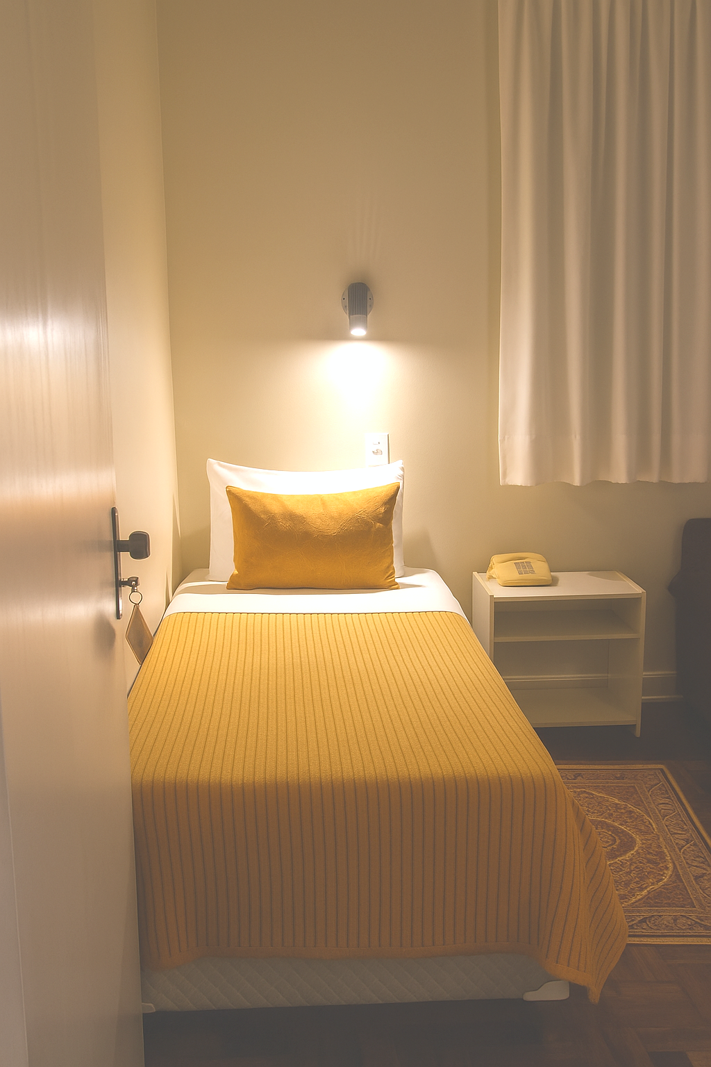 Quarto pequeno e aconchegante com uma cama de solteiro, um abajur com luz quente, cortinas brancas e uma mesa de cabeceira.