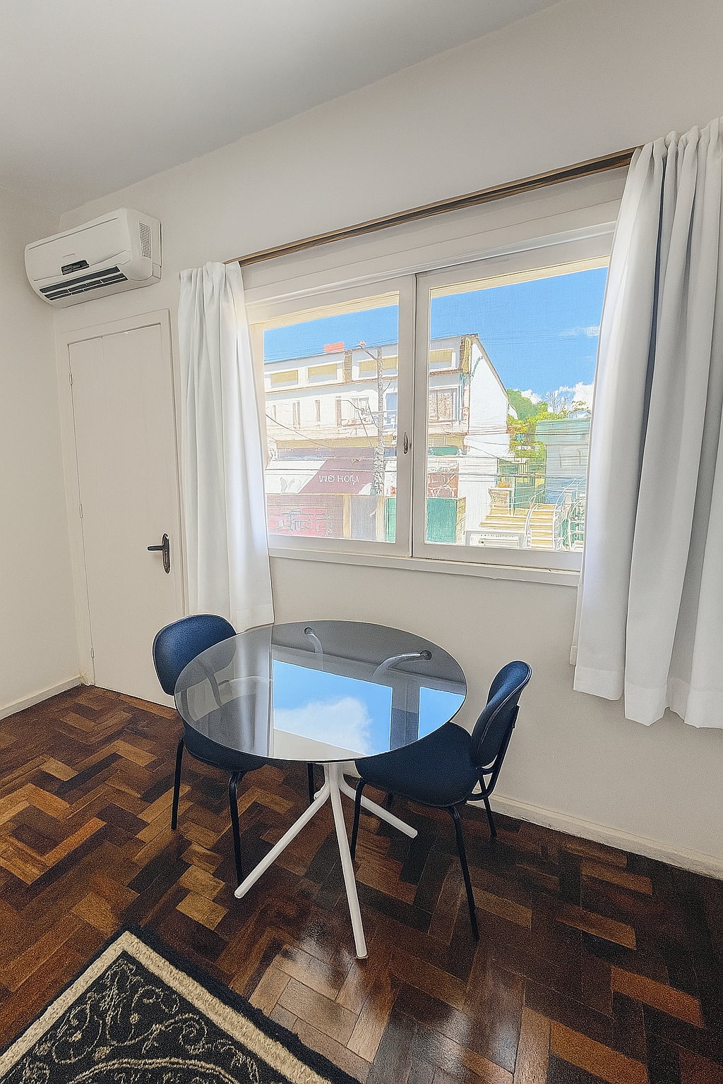 Sala iluminada com mesa redonda de vidro, duas cadeiras, cortinas brancas, janela e ar-condicionado de parede.