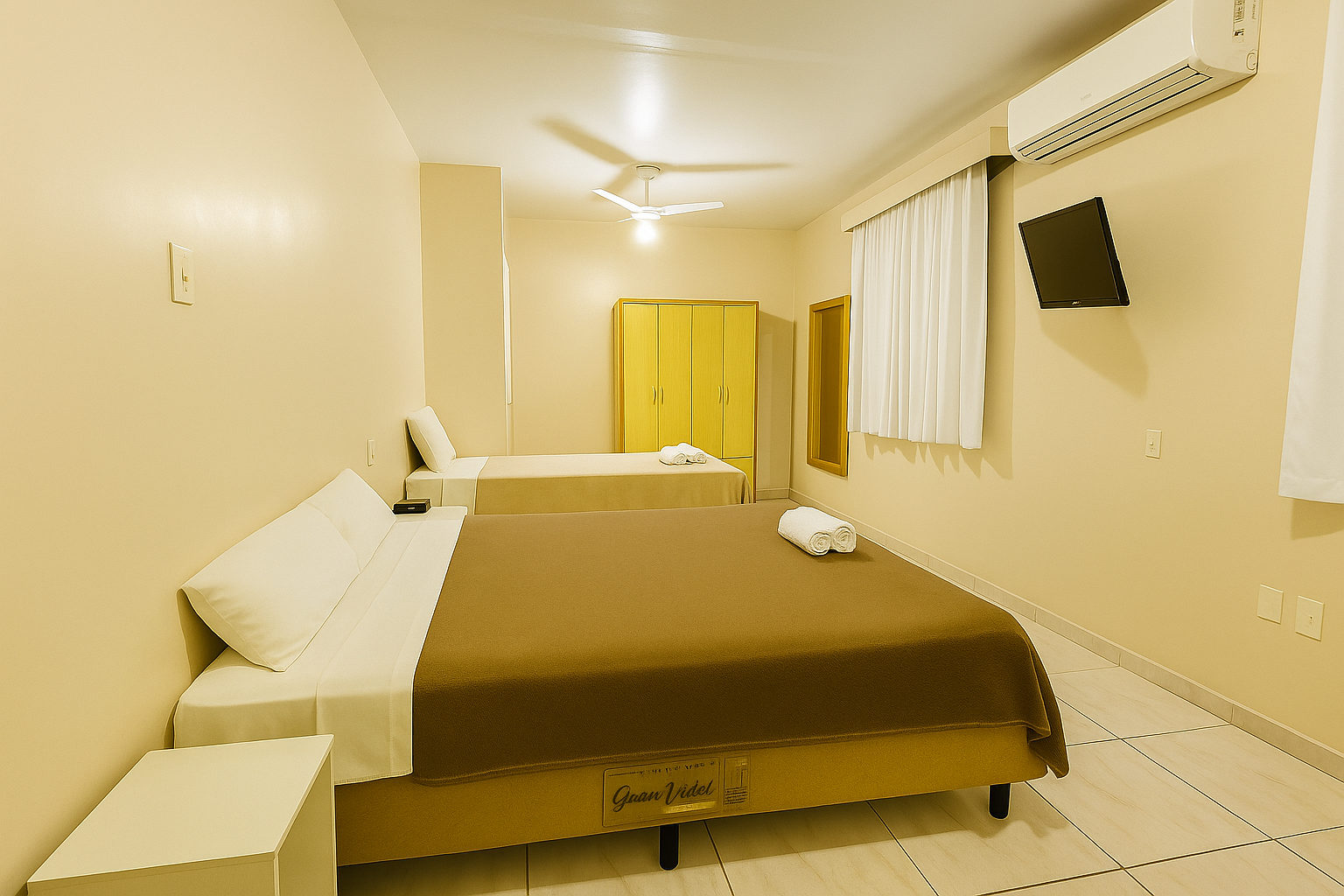 Quarto iluminado com cama, guarda-roupa, TV de parede e ventilador de teto em um ambiente bege.