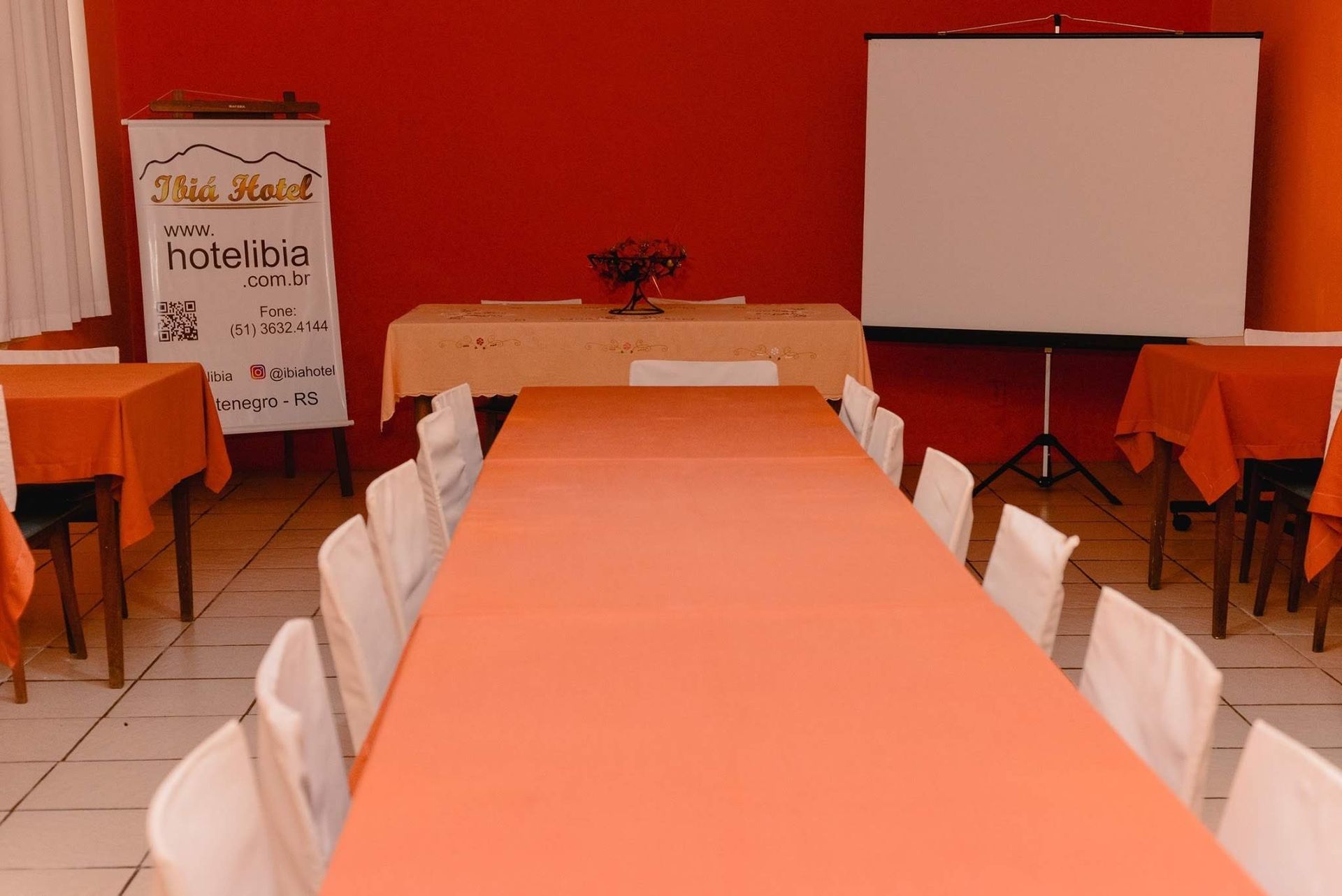 Sala de conferências com toalhas de mesa laranja, cadeiras brancas, tela de projeção e uma faixa à esquerda.