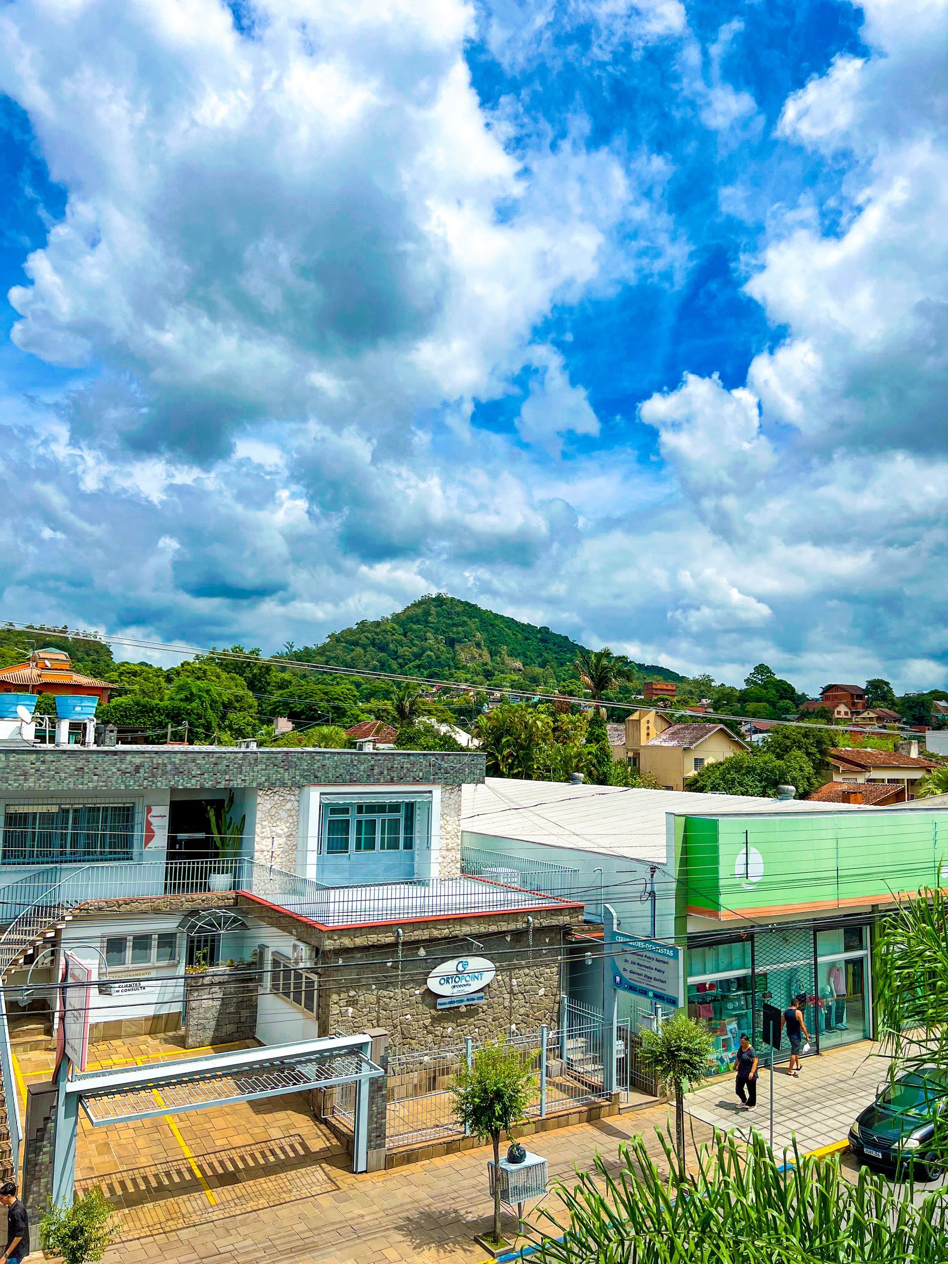 Cidade colorida na encosta de uma colina sob um céu azul brilhante com nuvens, apresentando edifícios baixos e uma colina verde ao fundo.