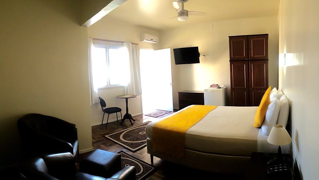 Quarto de hotel luminoso com cama, poltrona, TV, escrivaninha, janela e guarda-roupa.