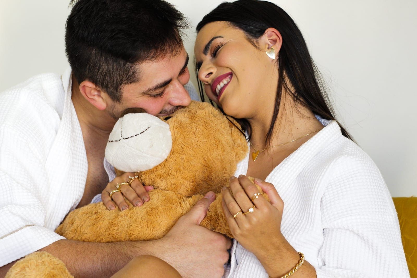 Casal se abraçando e sorrindo enquanto segura um ursinho de pelúcia na cama.