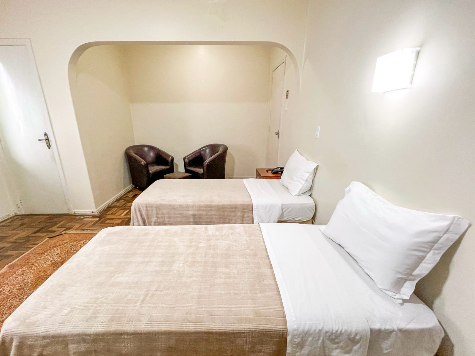 Quarto de hotel com duas camas de solteiro bem arrumadas, duas cadeiras e um aplique de parede em um interior branco simples.