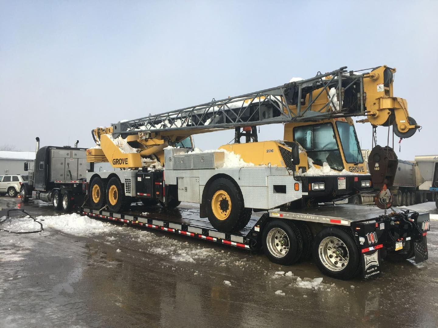 A large yellow and gray crane on a flatbed trailer, pulled by a black semi-truck, on a snowy surface.