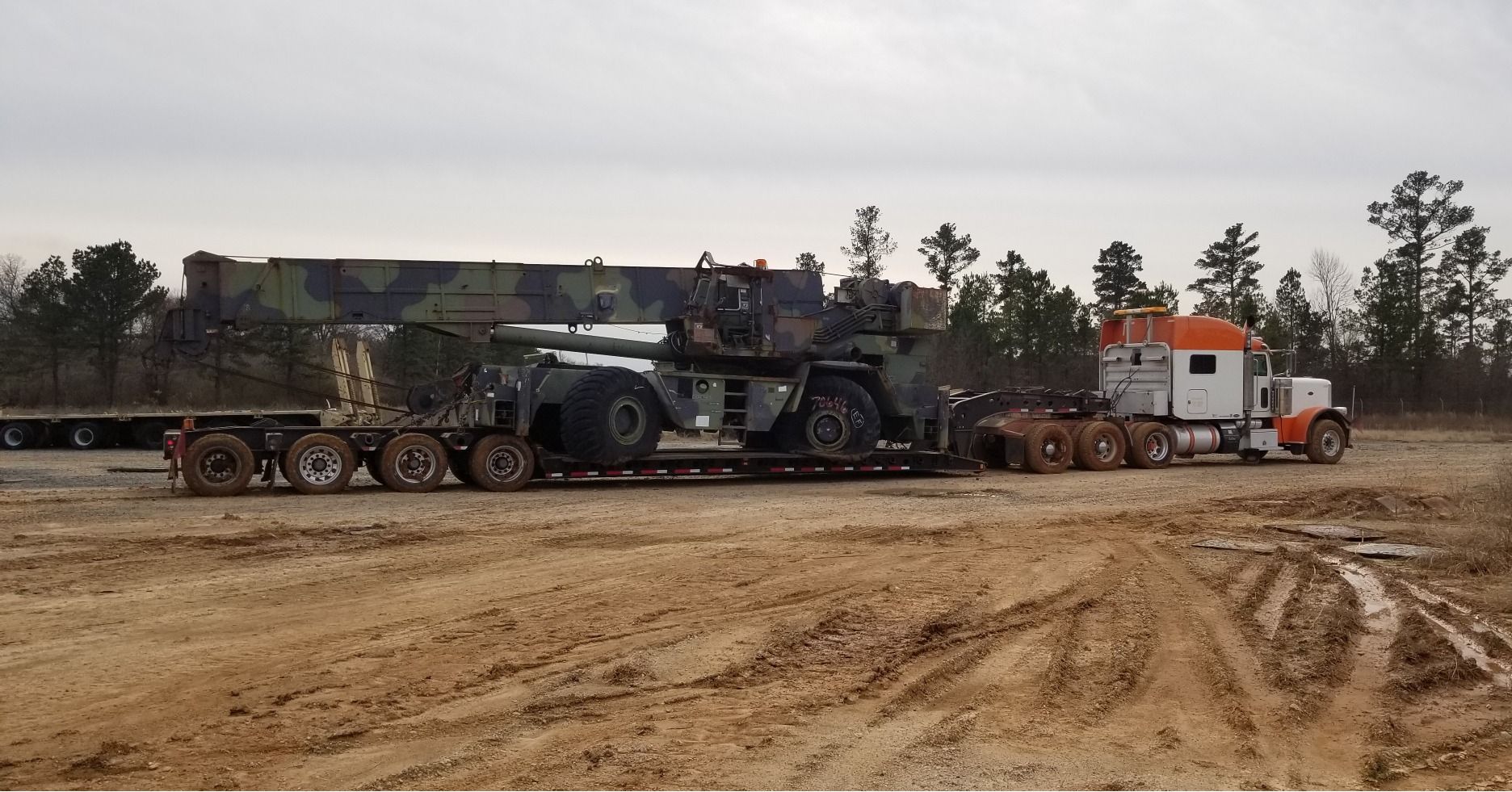 Heavy truck hauling a crane on a flatbed trailer across a dirt lot; trees in background.