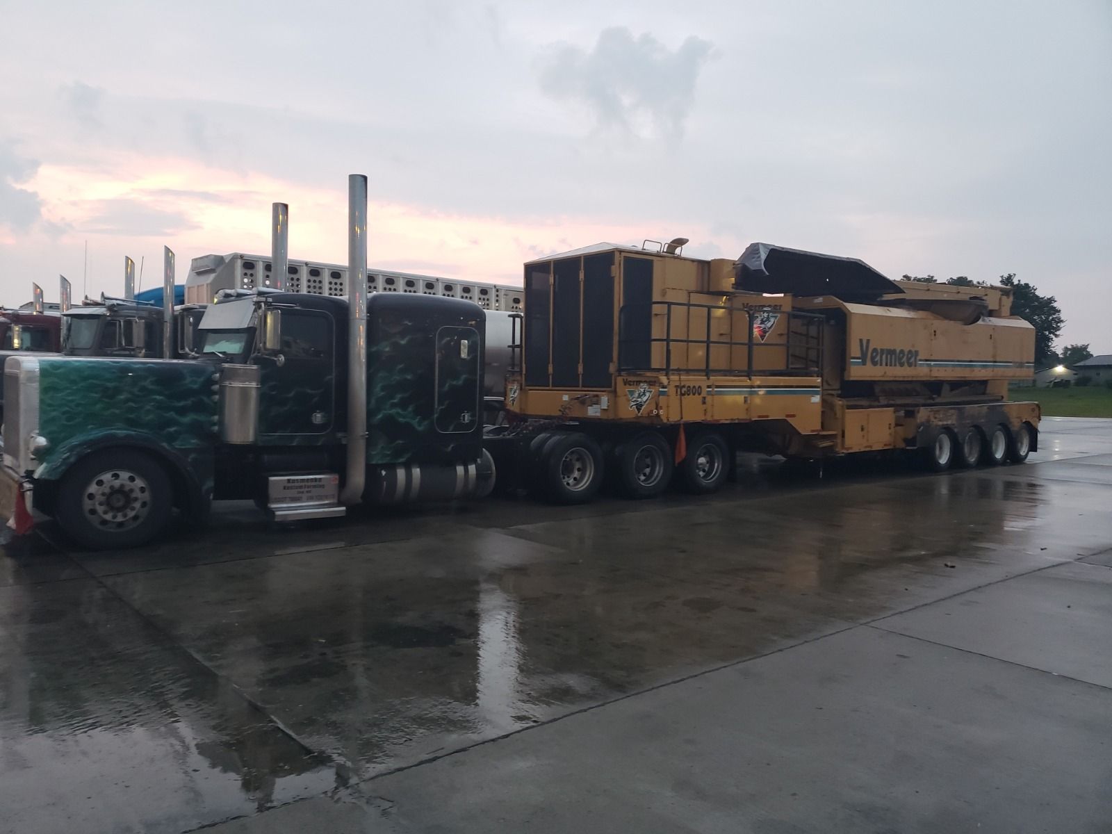 A large, black semi-truck hauls a yellow wood chipper on a trailer; parked on a wet surface.