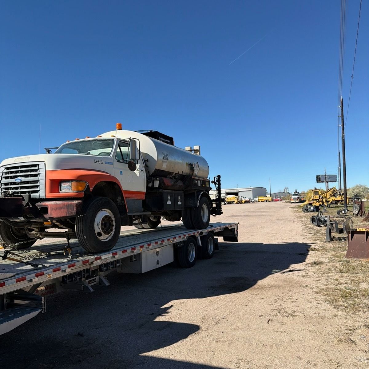 Water truck on a flatbed trailer on a gravel lot under a blue sky.