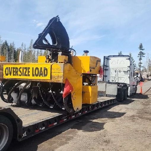 Yellow oversized snowblower on a flatbed trailer, being towed by a white truck on a road.