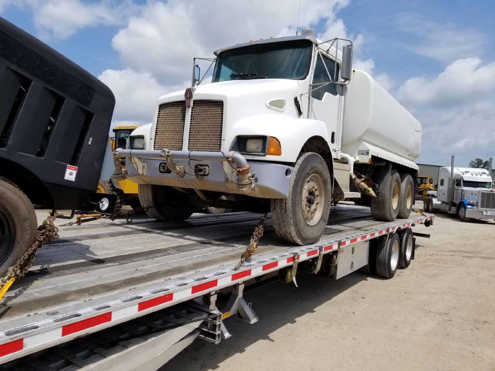 White dump truck loaded on a flatbed trailer in a dirt lot under a cloudy sky
