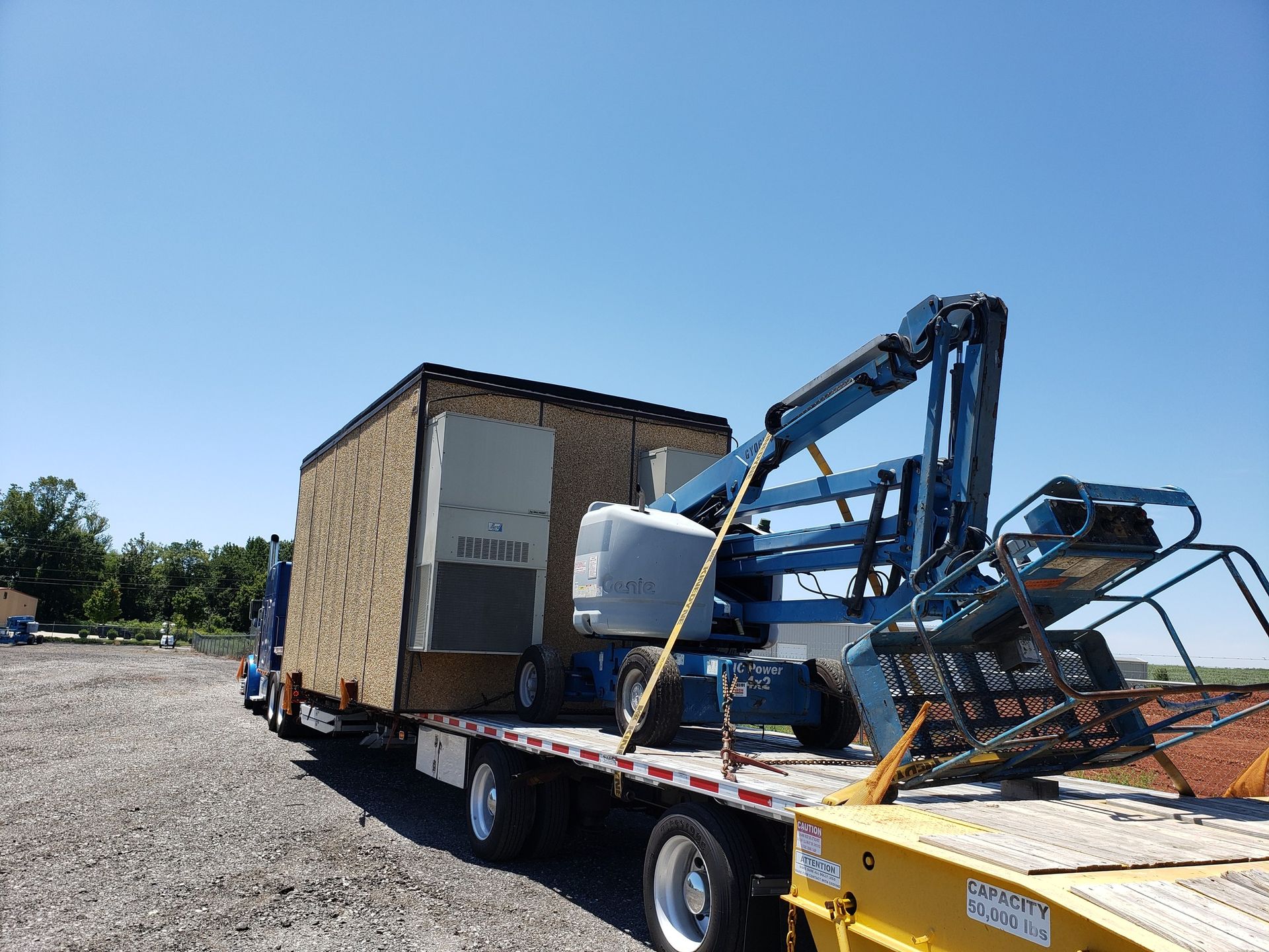 Flatbed truck carrying a large, wood-paneled structure and a blue lift against a blue sky.