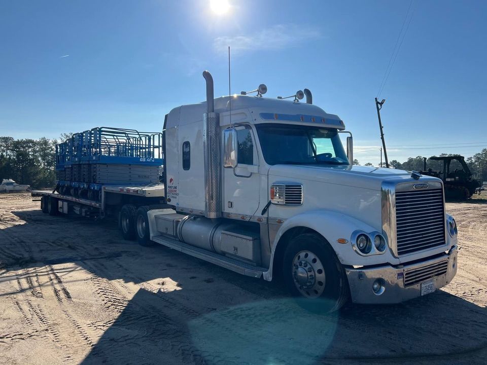 White semi-truck with flatbed trailer carrying blue metal frames on a sunny day in a dirt lot.