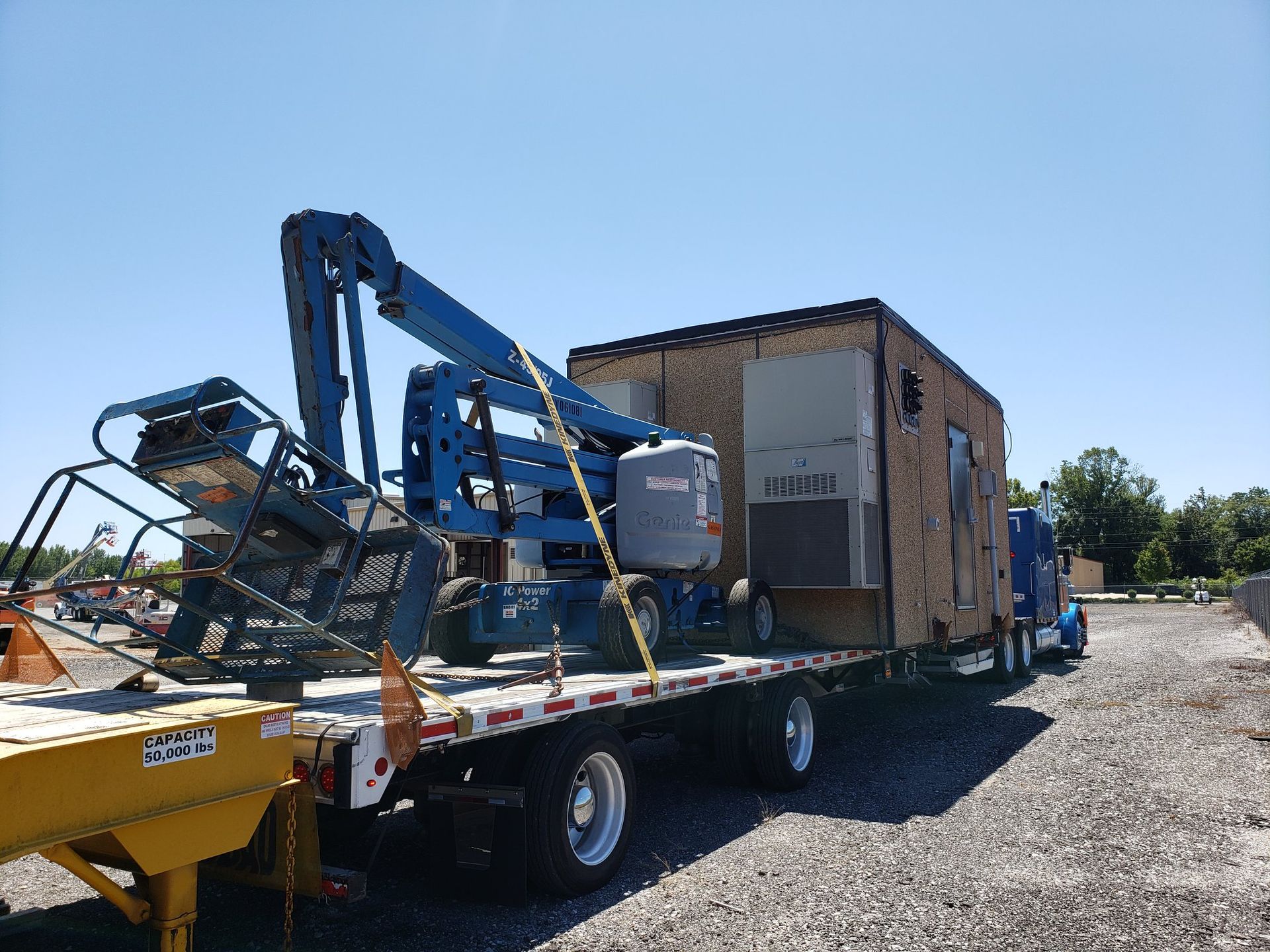Flatbed truck transporting a blue lift and a brown building under a bright blue sky.