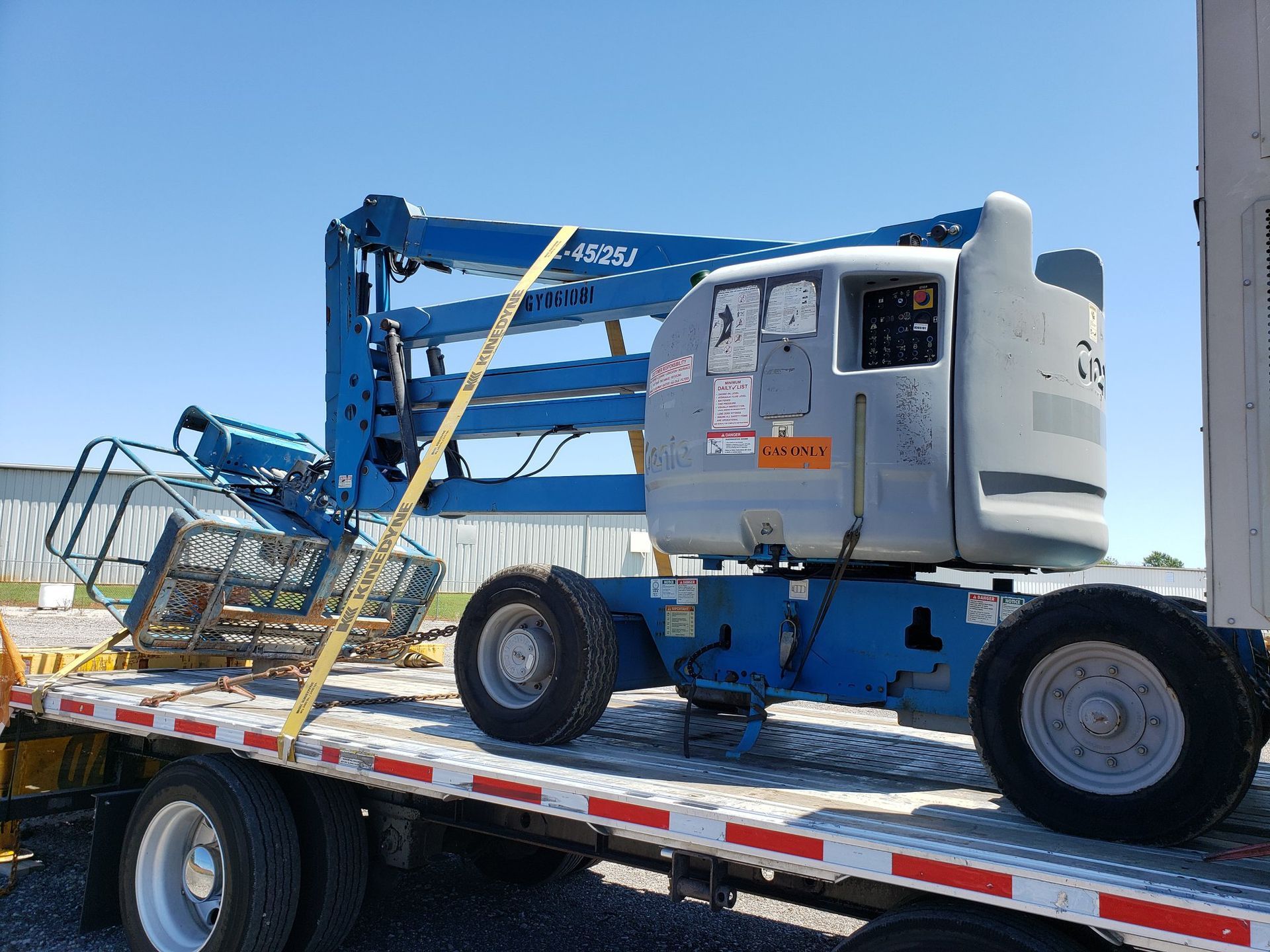 Blue aerial lift on a flatbed truck, secured with yellow straps, outdoors under a blue sky.